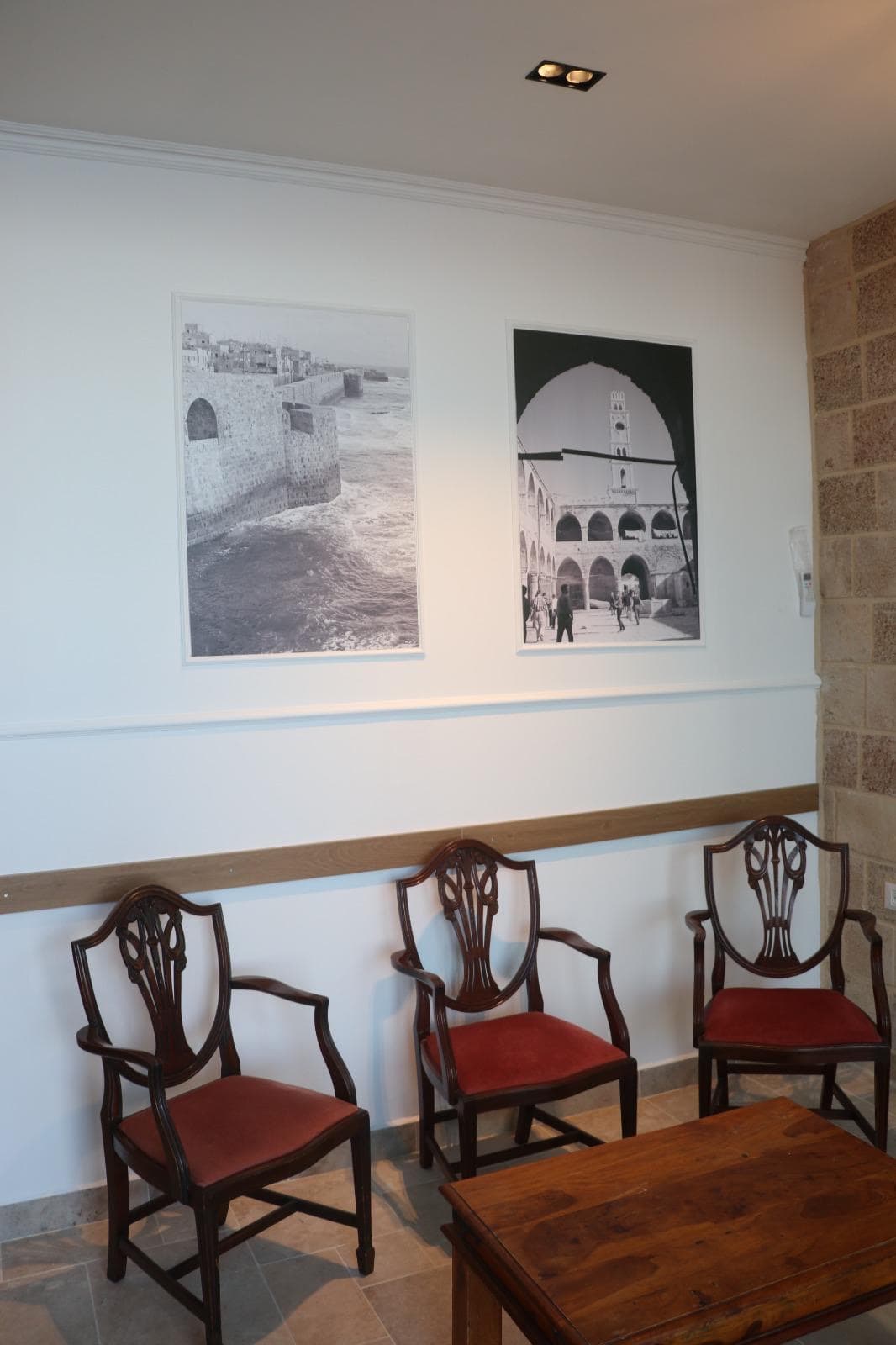 מסדרון עם שני תצלומים גדולים בגוני ספיה של הנמל העתיק והח'אן של עכו, מעל שלושה כיסאות עתיקים